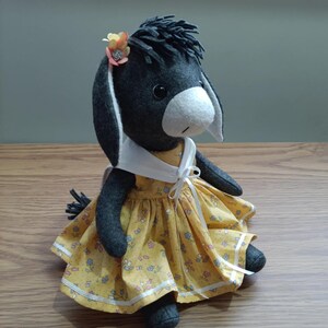 May include: A handcrafted felt donkey doll with dark gray fur, a white muzzle, and black eyes. The doll wears a yellow dress with a small floral pattern, a white sailor-style collar with a ribbon tie, and a small orange flower accessory on its head.