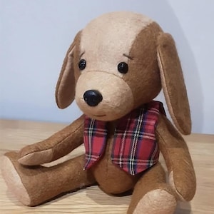 Op de afbeelding: Een pluche hondenspeeltje gemaakt van bruine en beige stof met lange slappe oren en zwarte kraaloogjes. De hond heeft een zwarte neus en draagt een rood, zwart en wit geruit vestje. Het speeltje zit in een zittende houding met de armen en benen naar voren.