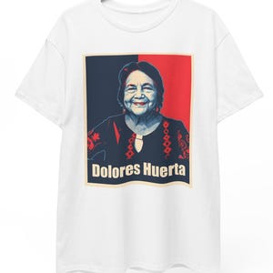 May include: White t-shirt featuring a portrait of Dolores Huerta. The portrait is set against a red and black background, with the name "Dolores Huerta" printed in a rectangular box at the bottom. The shirt is hanging on a wooden hanger.