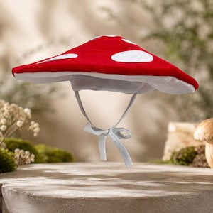 May include: A wide-brimmed hat shaped like a mushroom cap, featuring a vibrant red top with white circular patches and a white underside. The hat includes a white chin strap with fabric ties that hang down from the center of the brim.