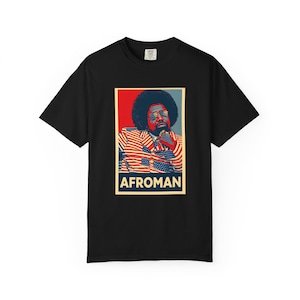 Puede incluir: Camiseta negra con un gráfico de un hombre con afro, gafas de sol y una chaqueta con un diseño de bandera estadounidense. La palabra "AFROMAN" está impresa en un recuadro rectangular en la parte inferior del gráfico.