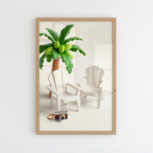 May include: A framed print featuring a miniature scene with two white Adirondack chairs, a palm tree with green coconuts, and a vintage camera. The artwork is set against a white background.