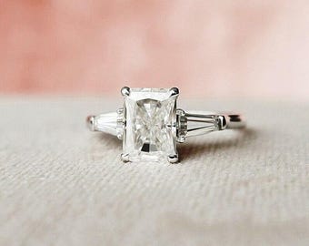 Pode incluir: Um anel de prata com um diamante de corte radiante, ladeado por dois diamantes baguette menores. O anel é colocado sobre um fundo rosa e branco suave. O diamante central é a característica principal.