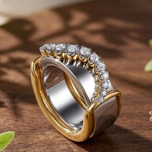 May include: A two-tone ring featuring a wide silver-colored band encased by a polished gold-colored frame. The top section of the ring is adorned with a curved row of small, clear, round-cut stones that follow the contour of the band's edge.
