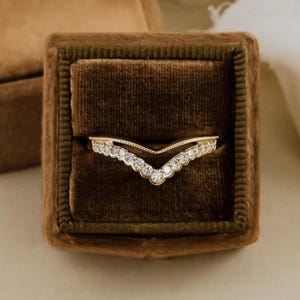 May include: A gold-colored, V-shaped ring with small, clear gemstones set along the top and bottom edges. The ring is displayed inside a brown velvet ring box, creating a luxurious presentation. The ring's design is delicate and elegant.