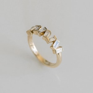 Könnte beinhalten: Ein Goldring mit einer Reihe von fünf klaren Baguette-Schliff-Steinen, die in einem versetzten Zickzack-Muster auf der oberen Hälfte des Rings angeordnet sind. Das Metall hat eine polierte Oberfläche und die Steine sind fest im Goldrahmen gefasst.