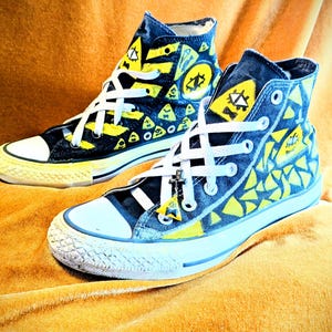 Op de afbeelding: Een paar hoge sneakers met een uniek ontwerp. De schoenen zijn marineblauw met gele geometrische patronen en witte veters. De zolen zijn wit en geel. Een kleine bedel hangt aan de veters. De sneakers staan op een goudgele ondergrond.