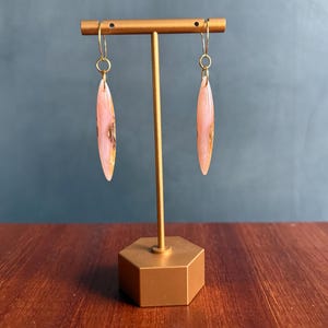 May include: A pair of dangle earrings with a teardrop shape. The earrings are a light pink color with gold and brown marbling. They hang from gold-colored ear wires and are displayed on a gold-colored earring stand.