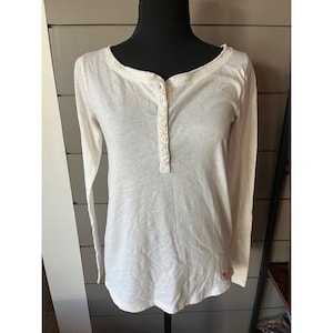 May include: Cream-coloured long-sleeve shirt with a Henley neckline and a row of buttons down the front. The shirt has a scoop neck and a small pink heart detail near the hem. The fabric has small pink flecks.
