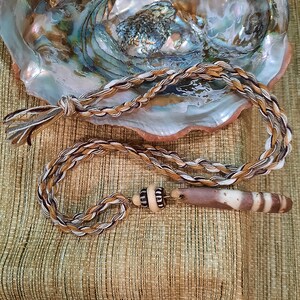 May include: A braided cord necklace with a brown and white striped pendant and a brown and white bead. The necklace is lying on a brown and white patterned surface.