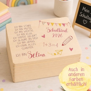 Könnte beinhalten: Eine rechteckige, helle Holzbox mit Klappdeckel, bedruckt mit deutschem Text, dem Namen Selina, der Jahreszahl 2026, einer Bleistiftzeichnung, einem Papierflieger, Herzen sowie einer Girlande aus bunten Wimpeln auf dem Deckel.
