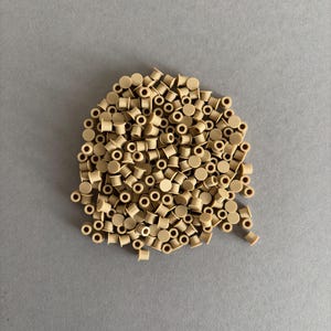 May include: A pile of small, beige plastic components with a cylindrical shape and a hole through the center. The components are scattered on a gray surface, creating a textured appearance. The components are uniform in size and color.