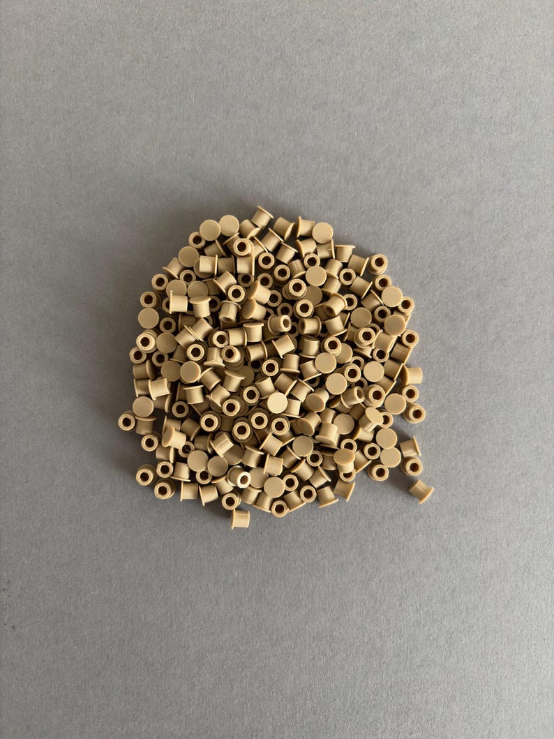 May include: A pile of small, beige plastic components with a cylindrical shape and a hole through the center. The components are scattered on a gray surface, creating a textured appearance. The components are uniform in size and color.