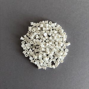 May include: A pile of small, white plastic pieces, each with a cylindrical body and a flat top, are scattered on a gray surface. The pieces appear to be uniform in size and shape, creating a textured appearance.