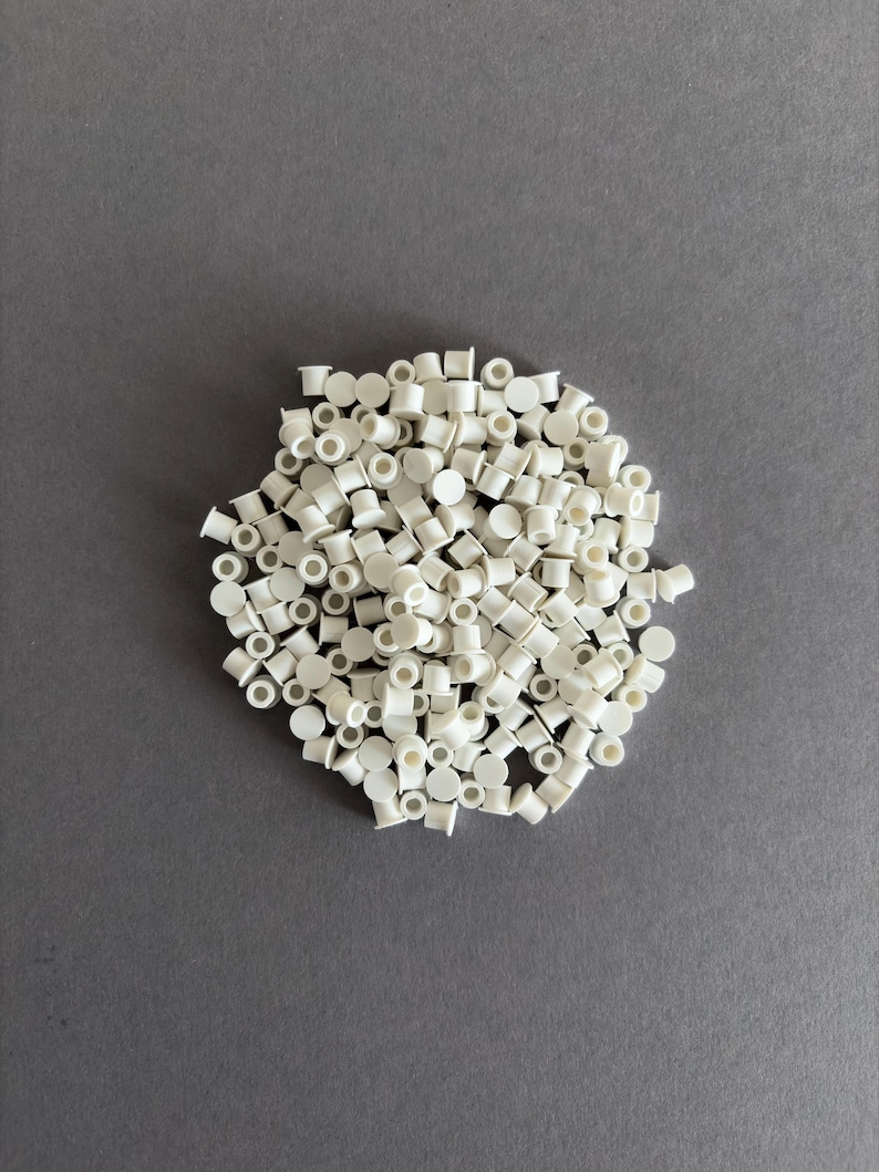 May include: A pile of small, white plastic pieces, each with a cylindrical body and a flat top, are scattered on a gray surface. The pieces appear to be uniform in size and shape, creating a textured appearance.