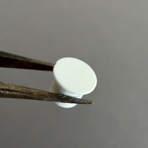 May include: A small, white, cylindrical object with a flat, circular top and a threaded base, held by a pair of tweezers. The object appears to be made of plastic or a similar material. The background is a solid, neutral gray.