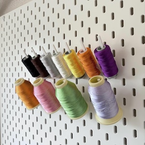 Puede incluir: Una colección de coloridas bobinas de hilo, incluyendo negro, marrón, gris, blanco, amarillo, naranja, morado, rosa, verde y lavanda. Las bobinas están dispuestas en un panel perforado blanco, mostrando una variedad de colores de costura y manualidades.