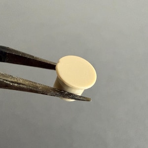 May include: A small, off-white plug held by tweezers. The plug has a flat, circular top and a cylindrical body with a ridged texture. The tweezers are dark metal, and the background is a solid, neutral gray.