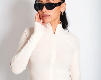 May include: Black sunglasses with a rectangular frame and gold accents. The sunglasses are worn with a cream-colored, long-sleeved top with a zipper. The model is wearing gold hoop earrings.