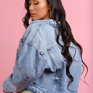 May include: Light blue denim jacket with distressed detailing. The jacket has a classic collar, button closures, and a relaxed fit. The back of the jacket features a ripped design. The model is wearing a beige skirt. The background is pink.