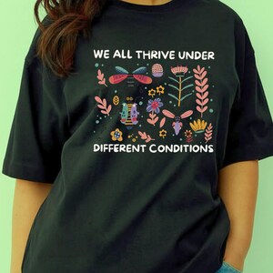 以下が含まれることがあります： 「WE ALL THRIVE UNDER DIFFERENT CONDITIONS」の白文字が入った黒いTシャツ。赤、ピンク、青、黄色、緑の色合いで、カラフルな花と昆虫のデザインが施されています。