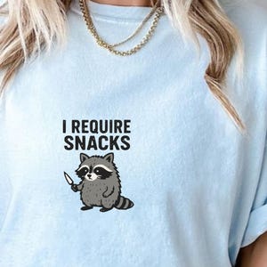 May include: Light blue t-shirt with the text "I REQUIRE SNACKS" above a cartoon raccoon holding a knife. The raccoon is gray, black, and white. The shirt is a solid light blue color.