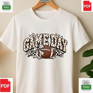 Może przedstawiać: Biała koszulka z krótkim rękawem z nadrukiem graficznym z napisem GAME DAY w czarno-białe kropki. Projekt zawiera brązową piłkę do futbolu amerykańskiego z białymi sznurowadłami, kokardę w kropki oraz małe akcenty w kształcie gwiazdek.