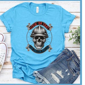 May include: A light blue t-shirt with a graphic of a skull wearing a hard hat. The design includes the text "Livin' on that Blue Collar Dolla" in red and blue. The shirt is paired with denim shorts.