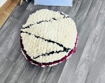 May include: A round, cream-colored Moroccan pouf with black and burgundy accents. The pouf is approximately 4.5 cm high and 20 cm in diameter. The design features black lines across the top and a burgundy border.