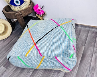 Può includere: Un cuscino da pavimento quadrato blu chiaro con un motivo a righe e linee geometriche colorate in rosa neon, verde, arancione e giallo. Il cuscino è su un pavimento in legno. Un cappello di paglia e un cestino intrecciato sono sullo sfondo.