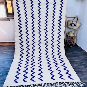 May include: A large, rectangular white rug with a repeating pattern of vertical, blue zigzags. The rug has a fringe along the bottom edge. The rug is made of a thick, woven material.
