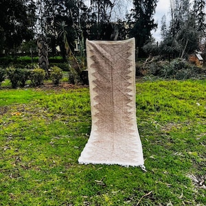 May include: A rectangular beige rug with a geometric pattern in shades of brown and cream. The rug is displayed outdoors on green grass, with a fringe along the bottom edge. The background features trees and foliage.
