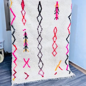 May include: A white, textured rug with vertical rows of geometric designs in black, pink, red, and yellow. The rug has a fringed edge and is displayed against a light blue wall and a gray floor.