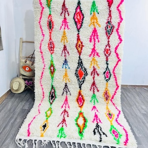 May include: A white, plush rug with a geometric pattern in vibrant colors. The design features diamond shapes and arrow-like motifs in pink, green, yellow, and red. The rug has a fringe along the bottom edge.