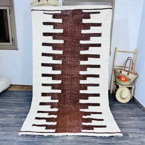 May include: A rectangular, off-white rug with a central, vertical band of dark brown geometric shapes. The rug features a repeating pattern of brown rectangles against a white background. The rug is displayed indoors.