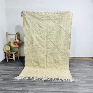 May include: A large, rectangular beige rug with a diamond pattern made of thick, light-colored yarn. The rug has a fringe along the bottom edge and is displayed against a white wall. A small wooden chair and a straw bag are visible in the background.