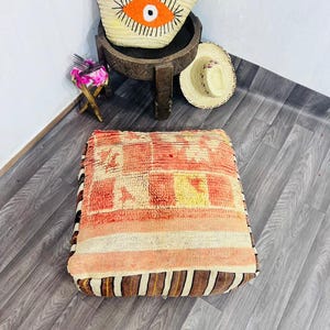 Op de afbeelding: Een vierkant vloerkussen met een patroon in perzik, oranje en crème. Het kussen heeft een gestreepte rand in bruin en crème. Een strooien tote bag met een oranje oogontwerp staat op een kleine houten tafel.