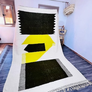 May include: A white rug with a geometric design featuring black and yellow shapes. The rug has a rectangular black shape at the top with a zig-zag edge, a yellow shape in the middle, and a black square at the bottom. The rug has a fringe border.