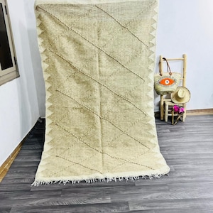 May include: A large, beige rug with a diamond pattern and fringed edges. The rug is made of a natural fiber and has a textured appearance. The design features a series of stitched lines forming the diamond shapes. The rug is displayed against a white wall.