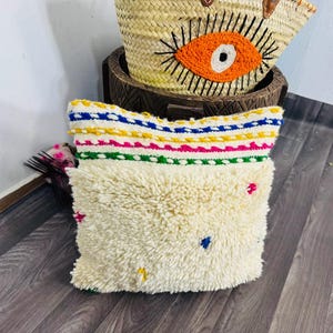 May include: A cream-colored, textured pillow with colorful embroidered details. The top edge features horizontal stripes in blue, yellow, pink, and green. Small, colorful embroidered accents are scattered across the pillow's surface. A woven basket with an eye design is in the background.