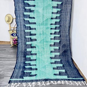 May include: A large, rectangular rug with a geometric pattern. The rug features a central turquoise design framed by a navy blue border with repeating rectangular shapes. White fringe trims the bottom edge. The rug is displayed against a white wall.