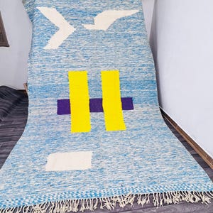 May include: A blue and white woven rug with a geometric design. The rug features a bird-like shape in white, two yellow rectangles, a purple rectangle, and a white rectangle. The rug has a fringed edge.