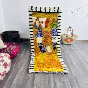 May include: A rectangular, hand-woven rug with a vibrant, abstract design. The rug features a yellow and orange central field with blue, green, and red accents. The borders are white with black stripes. A straw tote bag with an eye design is in the background.