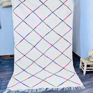 May include: A large, off-white rug with a colorful diamond pattern. The design features intersecting lines in blue, green, orange, and pink. The rug has a fringed edge and is displayed against a light blue wall.