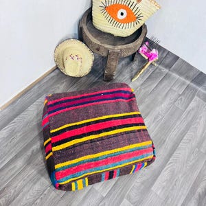 May include: A brown, square floor cushion with horizontal stripes in red, yellow, blue, and black. A straw tote bag with an orange eye design and a small straw hat are also visible.