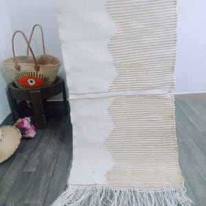May include: A cream-colored woven rug with a geometric design. The rug features a zigzag pattern in a lighter beige color and has a fringe at the bottom. A straw basket with leather handles and an evil eye design sits nearby.