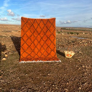 May include: An orange rug with a dark brown diamond pattern stands upright against a backdrop of a blue sky and arid landscape. A straw tote bag with a sun design sits nearby. The rug has a fringed edge.
