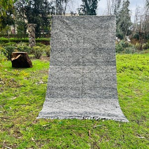 May include: A large, rectangular gray rug with a diamond pattern and fringe. The rug is displayed outdoors on a grassy lawn, with trees and a cloudy sky in the background. The rug's design features a repeating pattern of black lines.