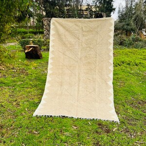 May include: A beige and white woven rug with a geometric diamond pattern and a fringed edge is displayed outdoors on green grass. The rug is hanging from a wooden structure.