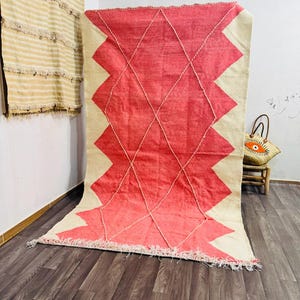 May include: A large, rectangular rug with a geometric design. The rug features a vibrant red zig-zag pattern against a cream-colored background. The design is accented with diamond-shaped stitching. The rug has a fringed edge and is displayed on a wooden floor.